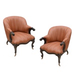 Par de poltronas em madeira lavrada com frisos e estilizações. Encostos em corbeille, ladeados por braços curtos rematados em volutas e apoiados em montantes arqueados. Assentos com frentes recortadas e saias sustentadas por pernas abauladas e recurvas. Pés pequenos assentados em rodinhas. Estofamento tacheado de cor tabaco. 87 x 71 x 67 cm.