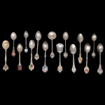 Conjunto de 15 colheres de metal prateado com decoração e materiais variados.  12 cm (maior). 9 cm (menor).