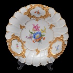 MEISSEN - Prato para doces em porcelana alemã de formato circular, ornamentado em esmaltes policromados e ouro. Borda moldada em gomos e recortada em lóbulos, ornada por pequenos adornos florais emoldurados em reservas com volutas e drapeados em alto relevo. Centro decorado por composição com flores do campo. Sob a base, marca da manufatura e numeração impressa. 30 cm.