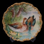 Prato para servir de porcelana francesa. Apresenta borda ondulada com friso em ouro. Centro com dois patos em lago e paisagem ao fundo. No verso marcado, Limoges. France. 34 cm.