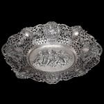 Fruteira baixa de formato ovalado, em prata possivelmente holandesa, repuxada, cinzelada e vazada. Borda larga e ondulada por friso de volutas, ornamentada por reservas com personagens, alternados por pequenos medalhões ovais, sobre fundo floral. Fundo apresentando quatro putti nadando com peixes em alto relevo. Marcas do teor de 800 mls. 6 x 36 x 27 cm. Peso: 565 gramas.