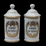 Par de Potes de farmácia em porcelana francesa de formato cilíndrico, com tampa cônica rematada por bolota. Bojo moldado com filetes dourados, ornamentado em policromia com guirlanda floral. Traz a inscrição: ESPRIT DEVENUS. França. Séc. XX. 27 cm.
