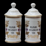 Par de Potes de farmácia em porcelana francesa de formato cilíndrico, com tampa cônica rematada por bolota. Bojo moldado com filetes dourados, ornamentado em policromia com guirlanda floral. Traz a inscrição ELIXIR DE LONGUE VIE. França. Séc. XX. 27,5 cm.