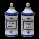 Par de Potes de farmácia em porcelana francesa de formato cilíndrico, com tampa cônica com larga faixa na cor azul, rematada por bolota. Bojo moldado com filetes dourados, ornamentado em policromia com guirlanda floral na cor azul e dourado.  Traz a inscrição: ALOES PURGAT E VISCUM ALBUM. França. Séc. XX. 26 cm.