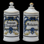 Par de Potes de farmácia em porcelana francesa de formato cilíndrico, com tampa cônica rematada por bolota. Bojo moldado com filetes dourados, ornamentado em policromia com guirlanda floral na cor azul e dourado. Traz a inscrição: RUSCUS ACULEATUS E BELLADONE. França. Séc. XX. 22 cm.