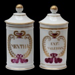 Par de Potes de farmácia em porcelana francesa de formato cilíndrico, com tampa cônica rematada por bolota em dourado. Bojo moldado com filetes dourados, ornamentado em policromia com guirlanda floral. Traz a inscrição: MENTHA E EXT:VALERIAN. 26 cm.