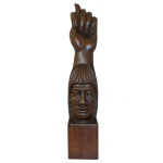 Escultura em madeira nobre representando cabeça de indígena brasileiro encimada por figa, amuleto popular para afastar o azar. Base retangular lisa. 91 x 18 x 18 cm. Apresenta risco e pequenos lascados.