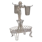 Paliteiro em prata brasileira repuxada e cinzelada, representando personagem carregando par de cestos, de pé sobre pedestal circular. Base com pequena salva oval bordada por galeria vazada, apoiada em pequenos pés recurvos. Pseudo contraste do Porto, usado no Brasil no séc. XIX e marca de prateiro não identificado de mesma época. 14  x 8 x 5 cm. Peso110 gramas.