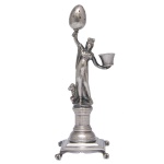 Paliteiro em prata portuguesa repuxada e cinzelada, no formato de divindade da antiguidade com leão aos seus pés. Apresenta-se erguendo ovo na mão direita e segurando recipiente na esquerda. Pedestal em coluna, ornado por frisos estilizados. Base quadrangular lisa com bordas em degraus, apoiada em pequenos pés de garra. Contraste desgastado do Porto do séc. XIX e marca de prateiro ilegível. 17 x 5 x 5 cm. Peso 171 gramas.