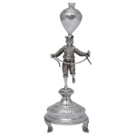 Paliteiro em prata europeia repuxada e cinzelada, representando funâmbulo equilibrando grande pião em sua testa e segurando corda. Apresenta-se ficando em um só pé sobre pedestal, apoiado em base circular almofadada. Pés recurvos e recortados em volutas. Contraste desgastado. 20 x 7 cm. Peso 139 gramas.