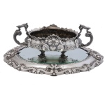 Centro de mesa oval com seu presentoir envidraçado em prata portuguesa repuxada e cinzelada, profusamente ornamentados em estilo D. João V com drapeados, reservas, volutas e folhagens. Bojo abaulado e baixo com borda ondulada, ladeado por largas alças em asas. Pés triangulares e recurvos. Prato com centro levemente elevado, ornado por borda acompanhando o estilo. Contraste da cidade do Porto Águia 833 mls e marca do prateiro. 13 x 37 x 19 cm / 1K199 gramas (floreira). 4 x 50 x 33 cm / 2K100 gramas (bandeja completa, com o vidro). Este lote encontra-se em Belo Horizonte, MG, e só estará disponível em nossa loja a partir do dia 13 de Outubro.