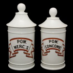 Par de potes de farmácia de porcelana francesa. Superfície branca apresentando rótulo com as inscrições: POM: MERC: S e POM: CONCOMB, envolto em moldura na forma de flâmula na cor vermelha. França. Séc. XX. 27 cm.