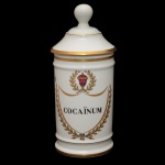 Pote de farmácia em porcelana francesa de formato cilíndrico, com tampa cônica rematada por bolota. Bojo moldado com frisos anelados e filetes dourados, ornamentado em policromia com guirlanda floral. Traz a inscrição Cocainum. Sob a base, marcas da manufatura de Limoges, France Au Pot de Gallien e Produits Dentaires Biodica. Séc. XX. 27 cm.