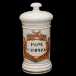 Pote de farmácia em porcelana da manufatura francesa. Superfície branca apresentando rótulo com a inscrição Pom: Campho envolta por composição policromada com motivos fitomórficos e guirlandas de flores e atributo mitológico grego, conhecido como a "Taça de Hígia". França. Séc. XIX. 26 cm.