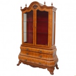 Bookcase estilo holandês em nogueira clara e radica. Alçada de formato retangular, com par de portas envidraçadas e interior com prateleiras em vidro sobre fundo em tecido de cor vinho. Frontão recortado em arcos ornados por frisos canelados e ponteiras nas laterais, centralizando adorno floral esculpido em alto relevo. Parte inferior formada por caixa bombée e contre bombée, apresentando três gavetões superpostos. Ilhargas acompanhando o movimento, alargando-se em direção à saia. Pés largos e recurvos rematados por garras. Ferragens em metal amarelo. 216 x 136 x 50 cm. Lote pertencente ao acervo de tradicional família paulistana de Américo Moreira dos Santos e Anella Mafalda Malzoni. Este lote encontra-se na residência em SP e a retirada devera ser feita a partir do endereço com a orientação da organizadora do leilão.