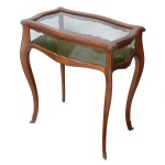 Mesa vitrine de formato retangular com laterais onduladas, em madeira lisa e lustrada, lavrada e ornada com frisos canelados. Tampo dobrável sobre caixa envidraçada, com fundo em tecido de cor verde musgo. Pernas curvilíneas rematadas por pequenos ornamentos em bronze dourado. Séc. XX. 74 x 65 x 39 cm. Lote pertencente ao acervo de tradicional família paulistana de Américo Moreira dos Santos e Anella Mafalda Malzoni.