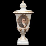 Urna com tampa em porcelana alemã, ornada com perolados e folhagens em relevo realçados a ouro. Bojo no formato de copo com borda revirada, ornamentado em esmaltes policromados apresentando busto de Ludwig von Beethoven em reserva. Tampa com larga pega semiesférica encimada por bolota. Base com pequena coluna sobre friso circular, apoiada em peanha quadrangular. Marca contemporânea da manufatura de Fraureuth e selo da Casa Castro  Shopping Ibirapuera  São Paulo. 59 x 28 cm. Apresenta restauro antigo. Lote pertencente ao acervo de tradicional família paulistana de Américo Moreira dos Santos e Anella Mafalda Malzoni.