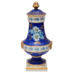 Urna com tampa em porcelana alemã, de formato balaústre, ornamentada em policromia e ouro com filetes, frisos florais e frente apresentando composição com folhagens e margaridas. Bojo no formato de cúpula, ladeado por pegas estilizadas e encimado por longo gargalo em corneta. Tampa abobadada com adorno piriforme. Pé circular esparramado sobre base quadrangular. Sob a base, marcas da manufatura RW  Bavaria  Made in Germany e selo da Casa Castro  Shopping Ibirapuera  São Paulo. 29 x 14 cm. Lote pertencente ao acervo de tradicional família paulistana de Américo Moreira dos Santos e Anella Mafalda Malzoni.