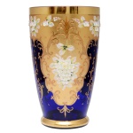 Floreira no formato de copo em vidro de cor Bleu du Roi, ricamente ornamentada em policromia por arranjos florais sobre fundo dourado distribuídos em painéis lobulados. Bordas lisas. 37 x 13 cm. Lote pertencente ao acervo de tradicional família paulistana de Américo Moreira dos Santos e Anella Mafalda Malzoni.