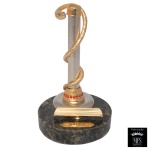 MFS  Porta caneta executado em liga metálica apoiada envolto em cobra de metal dourado. Base circular de bronze com pequenas pedras semipreciosas de cor vermelha. Apoio para a caneta e a placa de identificação Dentista. 15 x 9 cm. Peça nova sem uso. MFS tradicional fabricante de joias de elevado padrão de qualidade, reuniu técnica, talento e beleza artesanal. A experiencia e o pioneirismo no ramo joalheiro permitiram a criação de Produtos Finos para presentes que possuem componentes banhados a ouro 14 Quilates e as cores são resultado do aprimoramento da técnica revestida a fogo e da qualidade MFS. Lote pertencente ao acervo de tradicional família paulistana de Américo Moreira dos Santos e Anella Mafalda Malzoni.