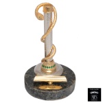 MFS  Porta caneta executado em liga metálica envolto em cobra de metal dourado. Base circular de bronze com pequenas pedras semipreciosas de cor verde. Apoio para a caneta e a placa de identificação escrito Medicina. 15 x 9 cm. Peça nova, sem uso. MFS tradicional fabricante de joias de elevado padrão de qualidade, reuniu técnica, talento e beleza artesanal. A experiencia e o pioneirismo no ramo joalheiro permitiram a criação de Produtos Finos para presentes que possuem componentes banhados a ouro 14 Quilates e as cores são resultado do aprimoramento da técnica revestida a fogo e da qualidade MFS. 15 x 9 cm. Lote pertencente ao acervo de tradicional família paulistana de Américo Moreira dos Santos e Anella Mafalda Malzoni.