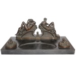 Tinteiro de mesa ornamentado por grupo escultórico em bronze patinado, apresentando casal de costas um para o outro, desnudos e deitados em assentos alinhados e apoiados em estrados realçados por volutas. Descanso de canetas escavado de formato ovalado, ladeado por recipientes embutidos com tampas circulares lavradas. Base retangular em granito preto. Assinado Michelangelo no verso da escultura. 22 x 50 x 25 cm. Pequeno bicado na base. Lote pertencente ao acervo de tradicional família paulistana de Américo Moreira dos Santos e Anella Mafalda Malzoni.