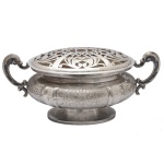 Centro de mesa com floreira em prata brasileira repuxada e cinzelada, ornamentado por painéis com arranjos florais gravados. Bojo globular achatado, ladeado por alças em S e encimado por grade vazada. Base circular no estilo com borda lisa. Contraste da prata brasileira 833 mls e marca de prateiro. 18 x 40 x 18 cm. 1k 854 gramas. Lote pertencente ao acervo de tradicional família paulistana de Américo Moreira dos Santos e Anella Mafalda Malzoni.