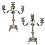 Par de candelabros para três velas de metal prateado. Coluna central com segmentos sustentando braços recurvos e bobeche central onde apresenta pássaros. base quadrangular com pés de garra. 37 x 29 cm. Lote pertencente ao acervo de tradicional família paulistana de Américo Moreira dos Santos e Anella Mafalda Malzoni.