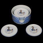 Conjunto de porcelana nacional para bebidas. Superficie de fundo branco com decoração com patos de laços azul. Marca da manufatura Porcelana Schmidt Brasil. Constando de 6 apoios para copos acondicionados em recipiente circular. apoio com pote para vinho (6 x 11 cm) e porta copos (9 x 6 cm). Lote pertencente ao acervo de tradicional família paulistana de Américo Moreira dos Santos e Anella Mafalda Malzoni.