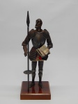 ESCULTURA, uma (1) representando Don Quixote, encontra-se de pé segurando um livro e uma lança, confeccionado em materiais diversos, base em madeira, medindo 26 cm altura.