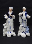 PORCELANA, par (2) esculturas representando homem e mulher, trajes de época, ambos segurando cesta de flores, base relevada em volutas e elementos vegetalistas na própria modelagem, pintadas a mão nas tonalidades azul e cor da pele sobre fundo branco, detalhes em rendilhados na técnica de barbotina (tecido de algodão tipo gaze embebido em massa líquida de porcelana que serve para moldar a renda na peça antes de ir ao forno), medindo em média 22 cm altura.