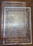 Duas Bandejas grandes confeccionadas em material  sintético transparente, sendo uma com acabamento  em metal  prateado. Medida Maior 52,5x39.