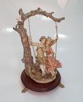 Belíssima escultura em resina italiana representando casal no balanço com base em madeira, sustentado por pés tripóides, medindo 43 cm x 26 cm.
