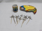 Lote composto de petisqueira na forma de peixe, copinho em porcelana e 11 espetos de petisco em metal prateado. Medindo a petisqueira 23cm x 19cm.