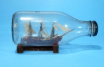 BARCO NA GARRAFA - Miniatura artesanal de veleiro policromado em marrom e vermelho, dentro de garrafa, suporte em madeira. 14 cm.