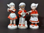 Lote contendo 03 (três) esculturas em porcelana coreana de tons rouge de fé e branco representando trio de crianças musicistas em trajes festivos típicos. Peças com pintura manual em ouro. Medem 14 cm de altura cada.
