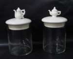 DIVERSOS (VIDRO E PORCELANA) - Par de potes para porta sachês de chá, confeccionados em vidro translucido, com tampas produzidas em porcelana esmaltada e vitrificada na cor branca encimadas por bules. Medidas alt total 16 cm x diam da boca 8 cm.