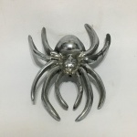 Adorno no formato de aranha em metal cinzelado e niquelado. Sinais de desgaste. Dimensões: 13 cm x 10 cm.