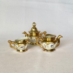 Conj. com três pequenas peças em porcelana, sendo: Uma cremeira e dois bules, um bule sem tampo, decorados com flores e dourado, med. 7cm de altura o maior considerando a tampa.