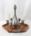 METAL PRATEADO - CP.X.MG COSTART - Diferenciada licoreira com seis cálices confeccionados em estanho, acompanha bandeja em madeira em formato sextavado guarnecida com o mesmo metal nas alças. Peças com manufaturada marcada em suas bases. Medidas: licoreira: 26,5 cm de altura total x 4 cm de diâmetro da borda/ cálices: 8 cm de altura x 4 cm de diâmetro da borda/ bandeja: 35,5 cm de comprimento total. Com sinais de uso.