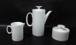 PORCELANA - Um bule para café, um açucareiro e uma cremeira em porcelana com esmaltagem branca, alças vazadas, pegas das tampas em formato de disco. Medidas: maior: alt total 18,5 cm x larg total 16,5 cm/ menor: alt total 8 cm x diam 9,7 cm.  Açucareiro e cremeira com bicados nas bordas.