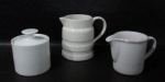 PORCELANA - Duas leiteiras, sendo uma da manufatura VERACRUZ (marcada no fundo), e um açucareiro em porcelana com esmaltagem branca, alças vazadas, pega da tampa em formato de disco. Medidas: maior: alt 12,5 cm x larg total 15 cm/ menor: alt 9 cm x larg total 14,5 cm. Uma leiteira com bicado na alça e tampa do açucareiro com bicado na borda.