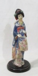ESCULTURAS - Escultura oriental em resina, ricamente detalhada e policromada, representando "Gueixa", apoiada sobre base circular em madeira patinada de preto. Med. alt 25 cm x diam da base 10,8 cm.