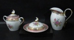 PORCELANA SCHMIDT - Uma leiteira, uma manteigueira e um açucareiro, em porcelana vitrificada branca, decorados com motivos florais em rica policromia ao centro, seguido de bordas adornadas com guirlandas de folhas e arabescos, generosos filetes e gregas em pintura à ouro, sobre faixa esmaltada na tonalidade rosa, pegas das tampas em forma estilizada e alças vazadas arrematadas com vasta douração, marcados no fundo. Med. maior: alt 14 cm x larg total 15,5 cm/ menor: alt total 8 cm x diam total 15,5 cm. Leiteira com alça colada, e manteigueira com ínfimo bicado na borda, conforme fotos extras.