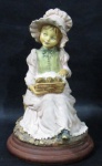 ESCULTURAS - Graciosa escultura em resina italiana, ricamente detalhada e policromada, representando "Menina sentada com cesto de passarinhos no colo", sobre base circular de madeira escalonada. Medidas alt 24 cm x diam da base 14 cm.