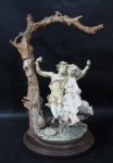 ESCULTURAS - Belíssima escultura em resina italiana, ricamente detalhada e policromada, representando "Jovem Casal no balanço", sobre base circular de madeira escalonada. Medidas alt total 40 cm x diam da base 22 cm. Falta a corda do balanço.