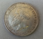 NUMISMÁTICA, Brasil, Império, uma (1) moeda, 2000 reis 1889, prata (900/916 milésimos), 3 sistema monetário, 4 tipo, 25,5gr, 37mm diâmetro, 2,7mm espessura, bordo serrilhado, 2 reinado, D. Pedro II, muito bom estado de conservação, anverso: Efígie de D. Pedro II, na orla a legenda PETRUS II D. G. C. IMP. ET PERP. BRAS. DEF. e o ano de cunhagem realizada à época na Província do Rio de Janeiro; reverso: no enxergo brasão do império na orla o dístico do valor de face e o DECRETO DE 1870.