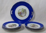 PORCELANA - REAL S. PAULO - Um prato para bolo com seis pratinhos em porcelana nacional vitrificada branca, galeria central apresentando cena galante em rica policromia, seguida de friso dourado, bordas ligeiramente onduladas e adornadas com larga faixa na tonalidade azul, finalizando com filetes em pintura à ouro, marcados no fundo, medindo: maior: 26 cm de diâmetro/ menor: 18 cm de diâmetro.