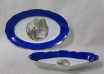 PORCELANA - REAL S. PAULO - Uma travessa e uma petisqueira ovaladas em porcelana nacional vitrificada branca, galeria central apresentando cena galante em rica policromia, seguida de friso dourado, bordas ligeiramente onduladas e adornadas com larga faixa na tonalidade azul, finalizando com filetes em pintura à ouro, marcadas no fundo. Med. travessa: comp 28 cm x larg 18 cm/ petisqueira: comp 22,5 cm larg 11,5 cm. Ambas possui bicados nas bordas.