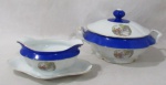 PORCELANA - REAL S. PAULO - Uma terrina e uma molheira com presentoir em porcelana nacional vitrificada branca, apresentando cena galante em rica policromia ao centro, seguida de friso dourado, bordas ligeiramente onduladas e adornadas com larga faixa na tonalidade azul, finalizando com filetes em pintura à ouro, pega da tampa em formato de flor, alças trabalhadas em folhas, marcadas no fundo. Med. terrina: alt total 16 cm x larg total 27,5 cm/ molheira; alt 9,5 cm x comp total 23 cm larg total 14,5 cm. Terrina possui pequenas falhas na douração.